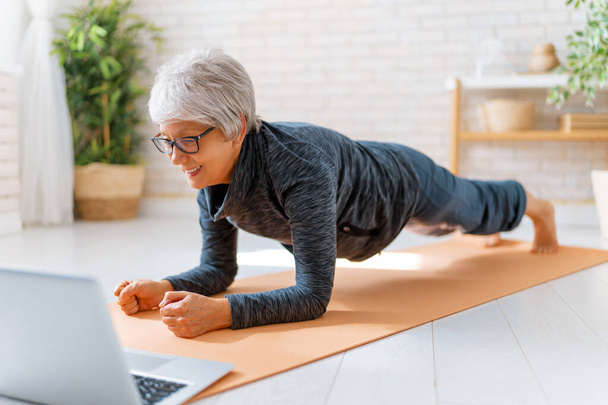 Senior woman in activewear watching online courses on laptop while exercising at home. - Fotografie, Obrázek