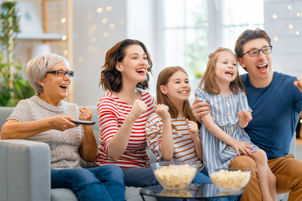 Щаслива сім'я дивиться телевізор з попкорном вдома. Бабуся, мати, батько і дочки проводять разом час.. - Фото, зображення
