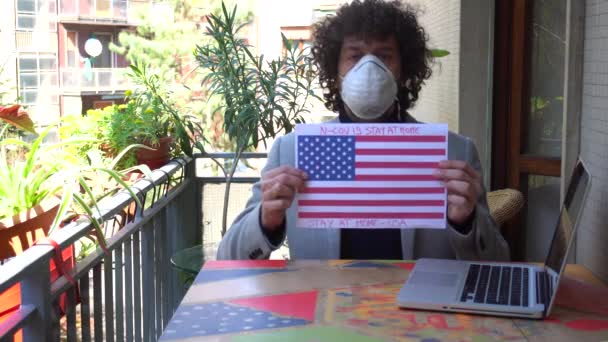 ヨーロッパ、イタリア、ミラノ-男性40歳自宅で自宅でn-cov19コロナウイルスの流行の検疫中にマスクをつけて-自宅で働いており、米国の旗が感染していることを示しています - 映像、動画