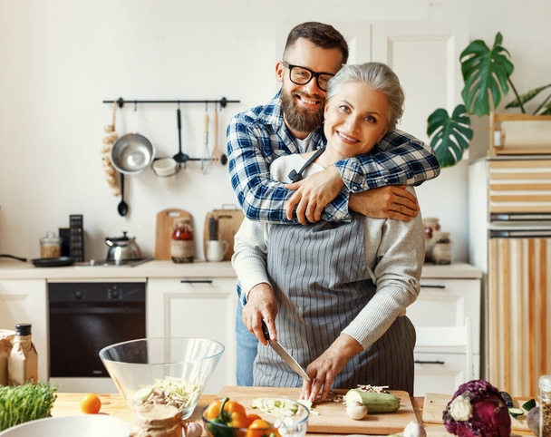 Veselý mladý vousatý muž objímající šťastnou zralou matku v zástěře při přípravě zdravého pokrmu s čerstvou zeleninou v domácí kuchyni - Fotografie, Obrázek