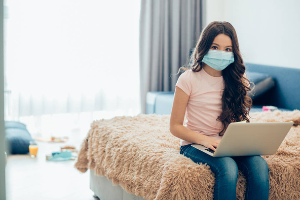 Calm long haired girl holding a modern laptop while sitting on a bed with a medical mask on her face. Template banner - Fotografie, Obrázek