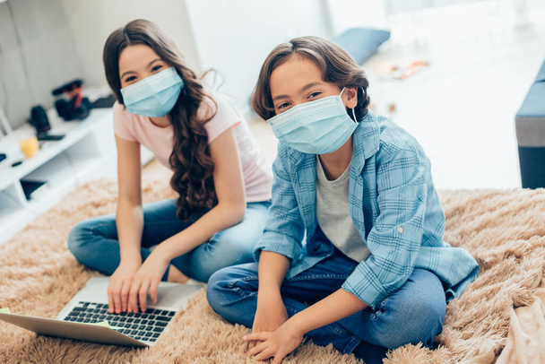 Girl and boy wearing medical masks at home and sitting together on the bed with a laptop in front of them - 写真・画像
