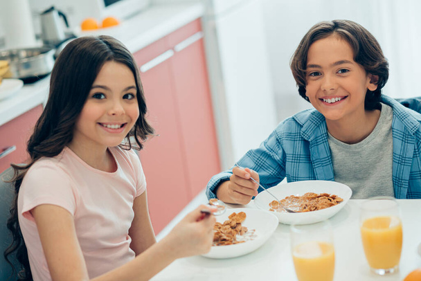 Contented girl and boy smiling while sitting together at home in their kitchen with bowls of tasty cornflakes with milk and glasses of juice - Photo, Image