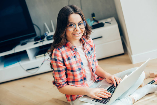 Positive pretty girl in casual clothes sitting on the floor with a modern laptop and smiling while putting hands on the keyboard - 写真・画像