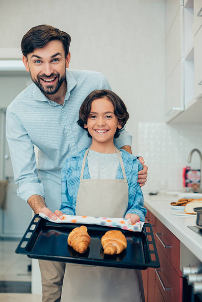 Contented boy holding a baking pan with two tasty croissants on it and smiling while a happy father standing next to him - 写真・画像