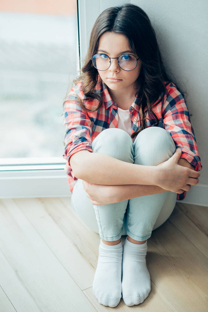 Calm girl in jeans and checkered shirt embracing her legs while sitting by the wall near the window - Фото, зображення