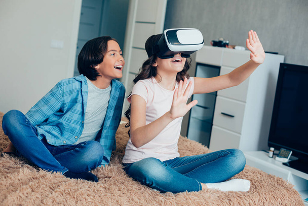 Positive boy smiling and watching his sister using virtual reality device and putting hands forward while touching something unreal - Zdjęcie, obraz