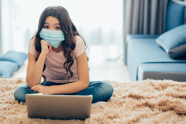 Cute girl in a medical mask putting her chin on a fist while sitting on the bed in front of a laptop in self isolation. Template banner - Photo, image