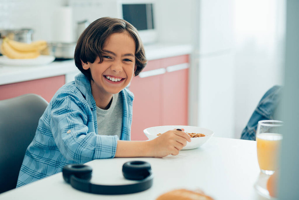 Funny kid sitting at the kitchen table with a spoon and a bowl of cereals - Photo, Image