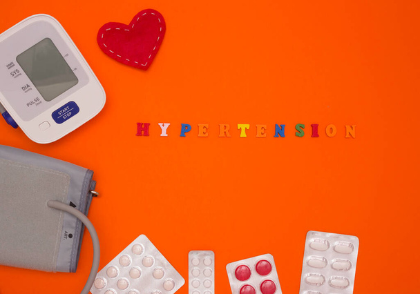 Pojem zdraví, krevní tlak. Slovo "hypertenze", elektronický monitor krevního tlaku, tablety a tkáňové srdce na oranžovém pozadí. Pohled shora.. - Fotografie, Obrázek