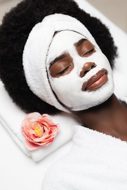 Портрет кудрявой африканской женщины с белой повязкой на голове и маской на лице, лежащей рядом с цветком - Фото, изображение