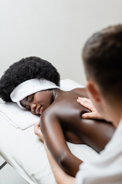 Masażysta masujący afrykańską kobietę w białej opasce na stole do masażu w salonie spa na niewyraźnym pierwszym planie - Zdjęcie, obraz
