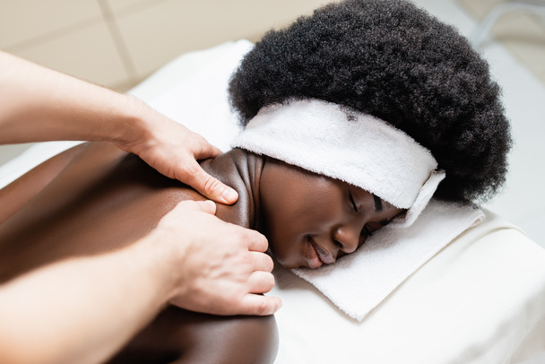 Close-up vista do massagista massageando mulher afro-americana na cabeça branca na mesa de massagem no salão de spa - Foto, Imagem