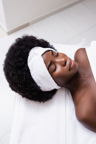 Bovenaanzicht van Afrikaanse Amerikaanse vrouw in witte hoofdband liggend op massagetafel in spa salon - Foto, afbeelding