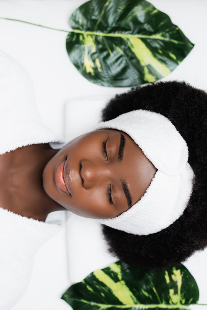 Вид сверху на африканскую женщину с белой повязкой на голове, отдыхающую рядом с зелеными листьями в спа-салоне - Фото, изображение