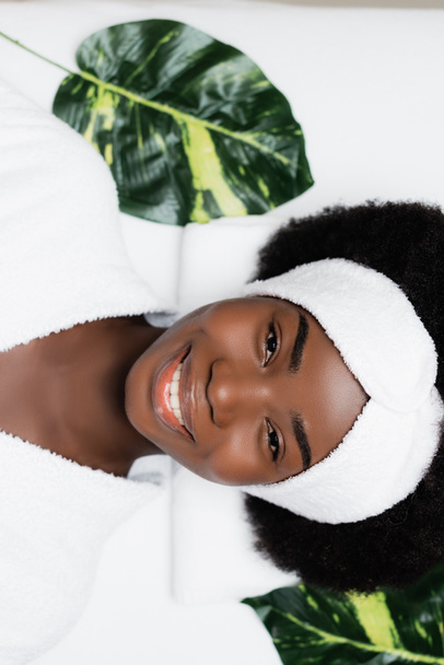 Vista superior de la mujer afroamericana feliz con diadema blanca mirando a la cámara, mientras descansa cerca de hojas verdes en el salón de spa - Foto, Imagen