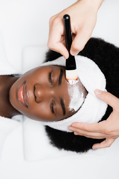 Spa terapisti, spa salonunda kozmetik fırçası olan Afrikalı Amerikalı bir kadının alnına yüz maskesi takıyor. - Fotoğraf, Görsel