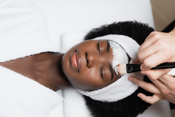 spa therapist applying face mask with cosmetic brush on forehead of african american woman wearing terry coat in spa salon - Foto, immagini