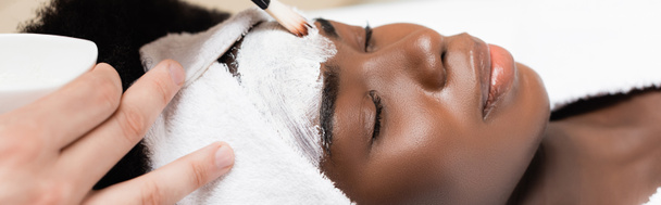 Close up άποψη του θεραπευτή spa εφαρμογή μάσκα προσώπου με καλλυντικά βούρτσα στο μέτωπο της αφρικανικής Αμερικής γυναίκα σε λευκό κεφαλόδεσμο, πανό - Φωτογραφία, εικόνα