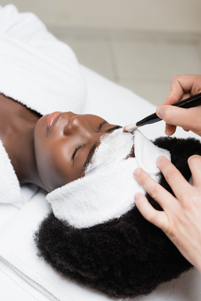 Bovenaanzicht van spa-therapeut aanbrengen gezichtsmasker met cosmetische borstel op het voorhoofd van Afrikaanse Amerikaanse vrouw in spa salon - Foto, afbeelding