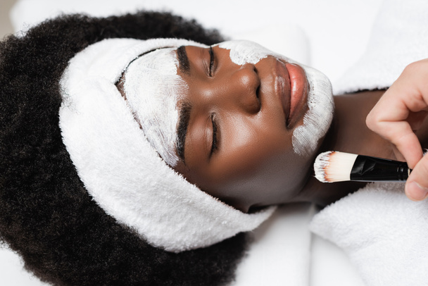 Vista de cerca de la mujer afroamericana positiva con los ojos cerrados acostado cerca de terapeuta de spa aplicando mascarilla en la barbilla en el salón de spa - Foto, imagen