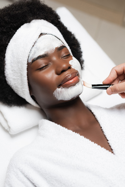 Mujer afro-americana positiva con los ojos cerrados acostada cerca de terapeuta de spa aplicando mascarilla en la barbilla en el salón de spa - Foto, Imagen