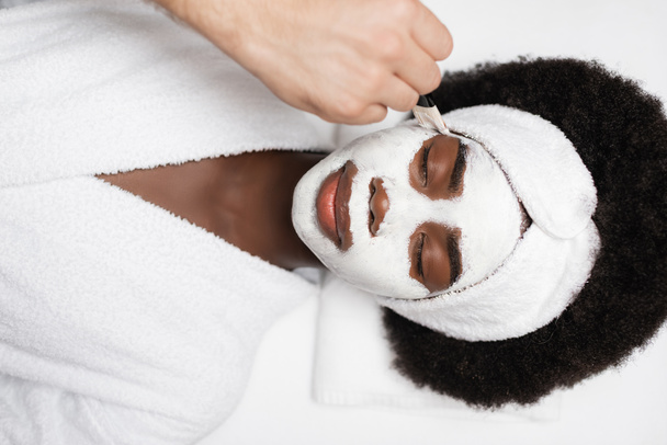 bovenaanzicht van positieve Afrikaan amerikaanse vrouw dragen badjas, liggen in de buurt spa therapeut aanbrengen gezichtsmasker op wang in spa salon - Foto, afbeelding