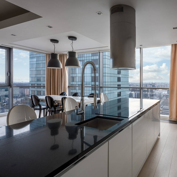 Luxury kitchen with black countertop and white cupboards open to dining area with big windows and city view - Foto, Bild