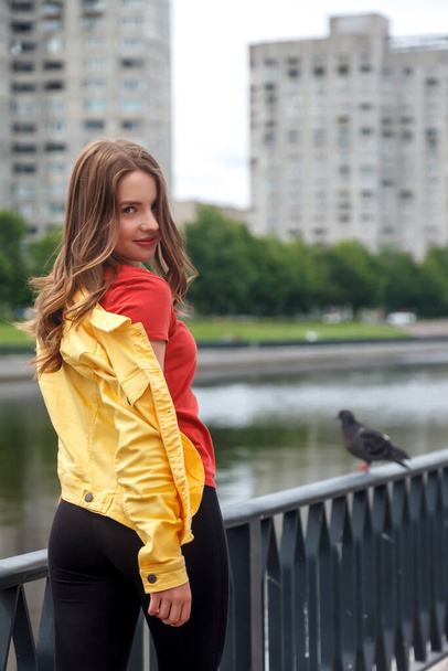 Pretty young sexy woman of athletic build in bright clothes on canal embankment in city. Walk in city center. Portrait of cute pretty female in yellow jacket and black pants on street - Фото, изображение