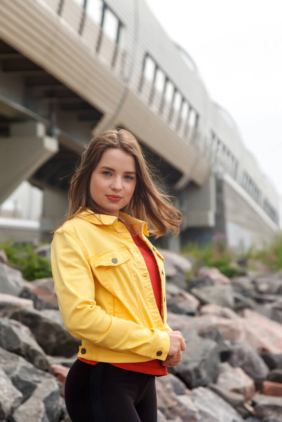 Pretty young sexy woman of athletic build is standing on stones on city beach. Walk through city center. Portrait of cute female in yellow jacket and black trousers on street - Foto, immagini