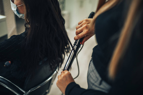 Γυναίκα με ιατρική μάσκα φτιάχνει τα μαλλιά της σε κομμωτήριο κατά τη διάρκεια πανδημίας.. - Φωτογραφία, εικόνα