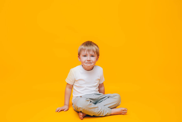Crying little boy rubs his eyes with his hands, a boy in a white t-shirt on a yellow background. - Photo, image