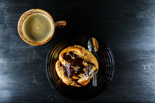 chocolate eclair on a plate and a cup of coffee on a black wooden background. View from above. - Φωτογραφία, εικόνα
