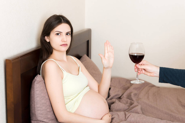 Yatakta kalan hamile kadın şarap içmeyi reddediyor ve cama jest yapmayı kesiyor. Hamilelik sırasında alkol yasak.. - Fotoğraf, Görsel