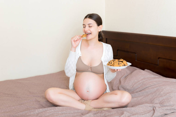 Pregnant woman enjoys eating cookies resting in bed at home. Unhealthy sweet pastry during pregnancy concept. - 写真・画像