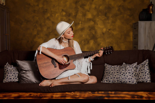 Beautiful young woman with guitar in interior of room. Cute woman on couch with guitar in her hands. Concept of home learning or playing guitar at home. Copyright space for site or banner or logo - Foto, Bild