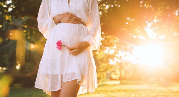 Beautiful photo of pregnant woman in white dress in sunlight of sunset at natural park. Close-up of pregnant woman with hands on her belly. Concept of pregnancy, maternity, expectation for baby birth. - Foto, afbeelding