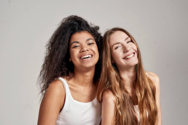 Portrait of two happy young diverse women in white shirts smiling at camera while posing together isolated over grey background - Foto, Imagen