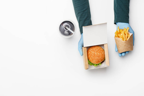Bestel in fast food restaurant. Ober in rubberen handschoenen geeft hamburger in kartonnen doos, en frites in papieren zak, en glas met cola, ijs en stro - Foto, afbeelding