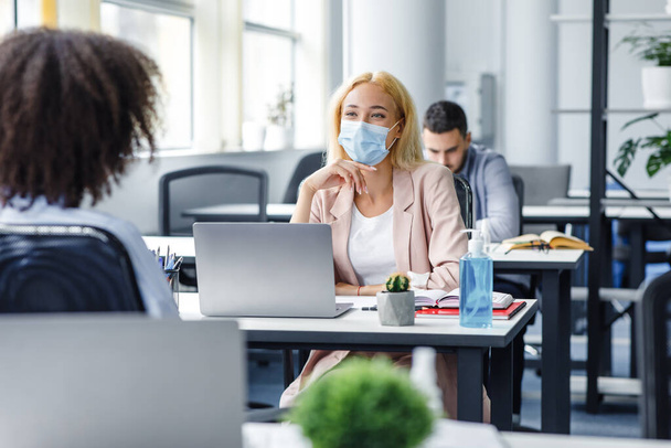 Communication of colleagues in office or client consultation and social distance. Millennial woman in protective mask talking with african american lady at workplace - Foto, Imagem