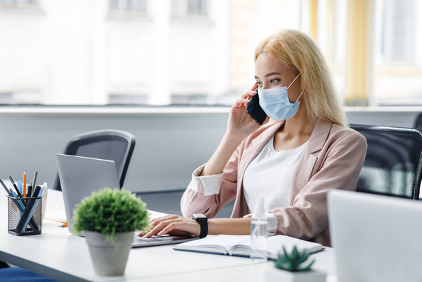 New normal and return to workplace after quarantine.Young attractive woman in a protective mask talks on smartphone and works in laptop, at table with antiseptic - Фото, изображение