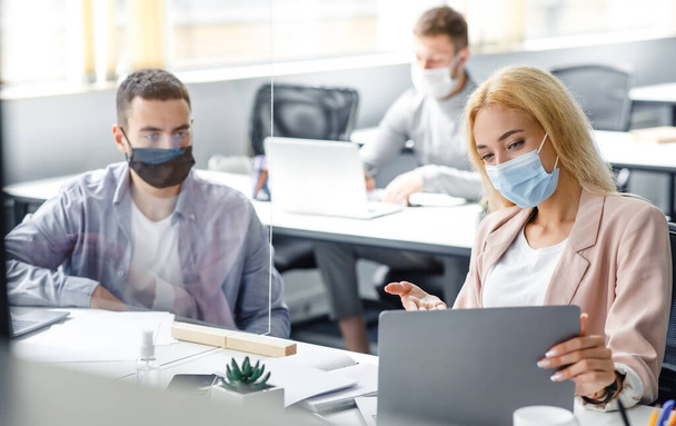 Meeting and discussing project during coronavirus epidemic. Young man and woman in protective masks look at laptop and communicate through glass board at workplace - Fotoğraf, Görsel