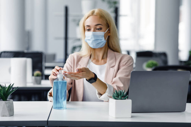Bezpieczeństwo i ochrona zdrowia w pracy w biurze. Millennial kobieta w masce ochronnej z inteligentnym zegarkiem używa antyseptyki w miejscu pracy z laptopem do dezynfekcji rąk - Zdjęcie, obraz
