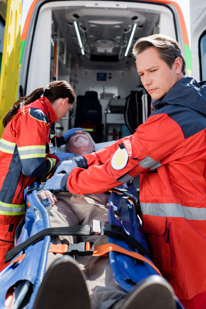 Selective focus of paramedics standing near elderly patient on stretcher and ambulance car - Photo, Image