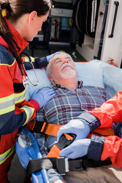 Enfoque selectivo del paciente que toca paramédico mientras colega cierra cinturones de camilla y coche de ambulancia - Foto, Imagen