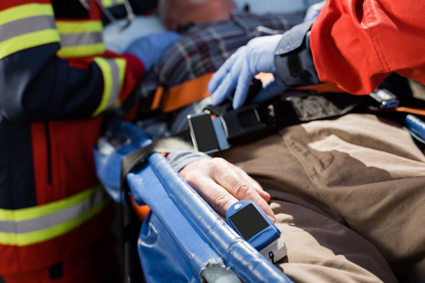 Vista recortada del paciente con monitor de frecuencia cardíaca acostado en camilla cerca de paramédicos  - Foto, Imagen