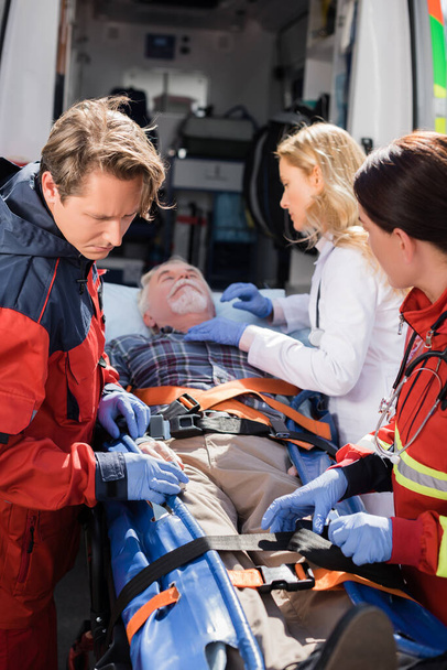Selective focus of paramedics and doctor standing near senior patient on stretcher and ambulance car - Photo, Image