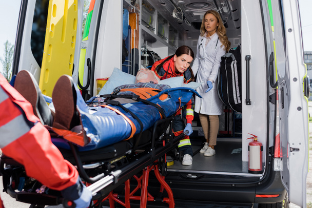 Enfoque selectivo del médico de pie cerca de los paramédicos que sostienen la camilla con el paciente en el coche de ambulancia  - Foto, Imagen