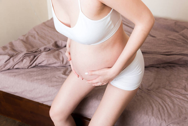 Młoda piękna kobieta w ciąży, w bólu, siedzi w łóżku w sypialni, przygotowując się do porodu. - Zdjęcie, obraz