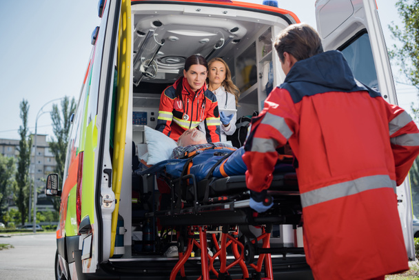 Concentration sélective du médecin avec tablette numérique debout dans la voiture d'ambulance près des ambulanciers avec patient sur civière à l'extérieur  - Photo, image